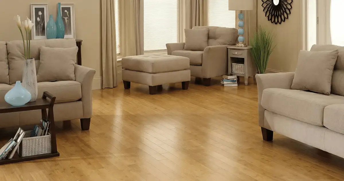 A living room with professionally installed bamboo flooring, beige sofas, a matching ottoman, a side table with a blue lamp, vases, framed pictures, and large windows with blinds and curtains in Cleveland, OH